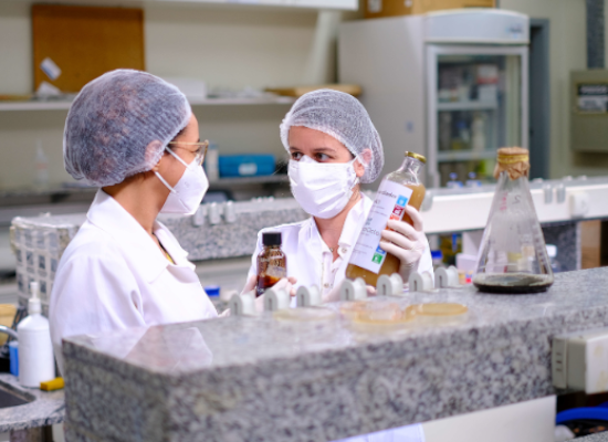 two women in protective clothes in a lab chatting with liquids in hand.