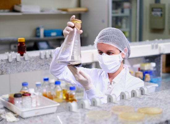 woman in protective clothes in a lab looking at a glass with liquid in it.