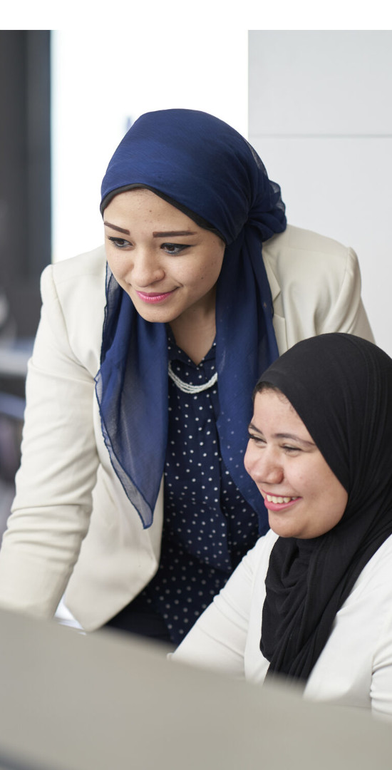 Two egypt women working on a computer. On is sitting while the other one is standing behind her.