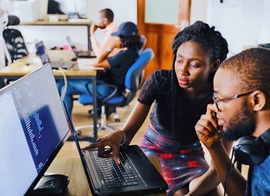  A teacher in a classroom showing to her student something on a laptop screen. A screen behind the laptop shows a programming language. 