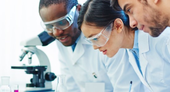 Three scientists with protective glasses and laboratory coats looking at samples in a laboratory.