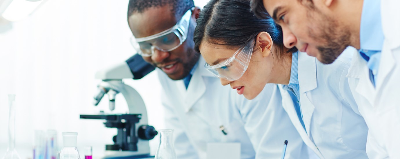 Three scientists with protective glasses and laboratory coats looking at samples in a laboratory.