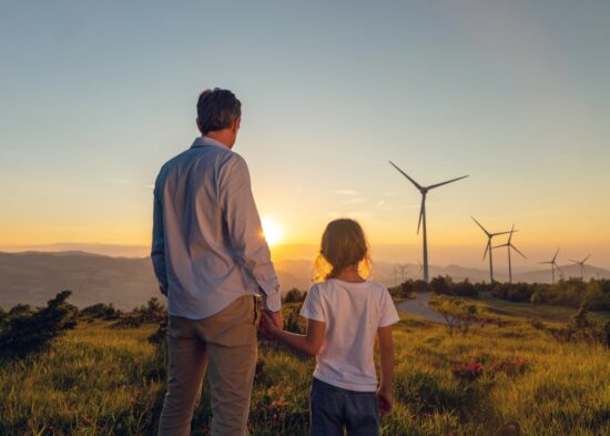  Father and daughter standing outdoors and looking towards a susnet scenery at a wind farm. 