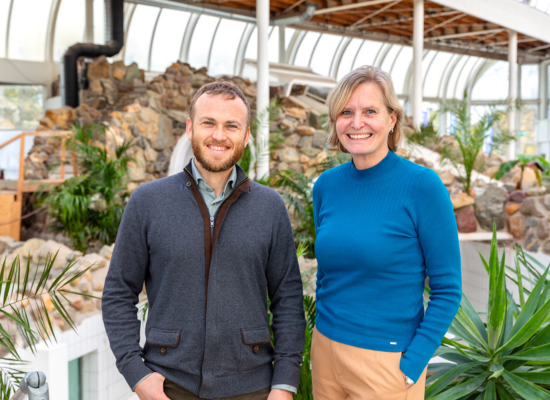 Blond man with beard next to a slightly taller blonde woman smiling into camera; background: some stone walls and farns, maybe a glass house