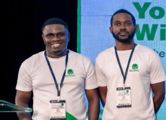 Two african men in white T-shirts with green logo smiling/ looking into camera, on a stage, exhibition pass on lanyard around their necks.